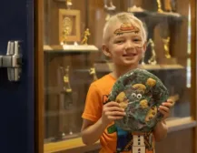 Smiling blond boy in an orange shirt holds a decorated round art piece, with trophy cabinets in a museum background.