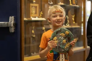Smiling blond boy in an orange shirt holds a decorated round art piece, with trophy cabinets in a museum background.