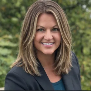 Professional headshot of a smiling woman with shoulder-length light brown hair, wearing a dark blazer outdoors with greenery in the background.